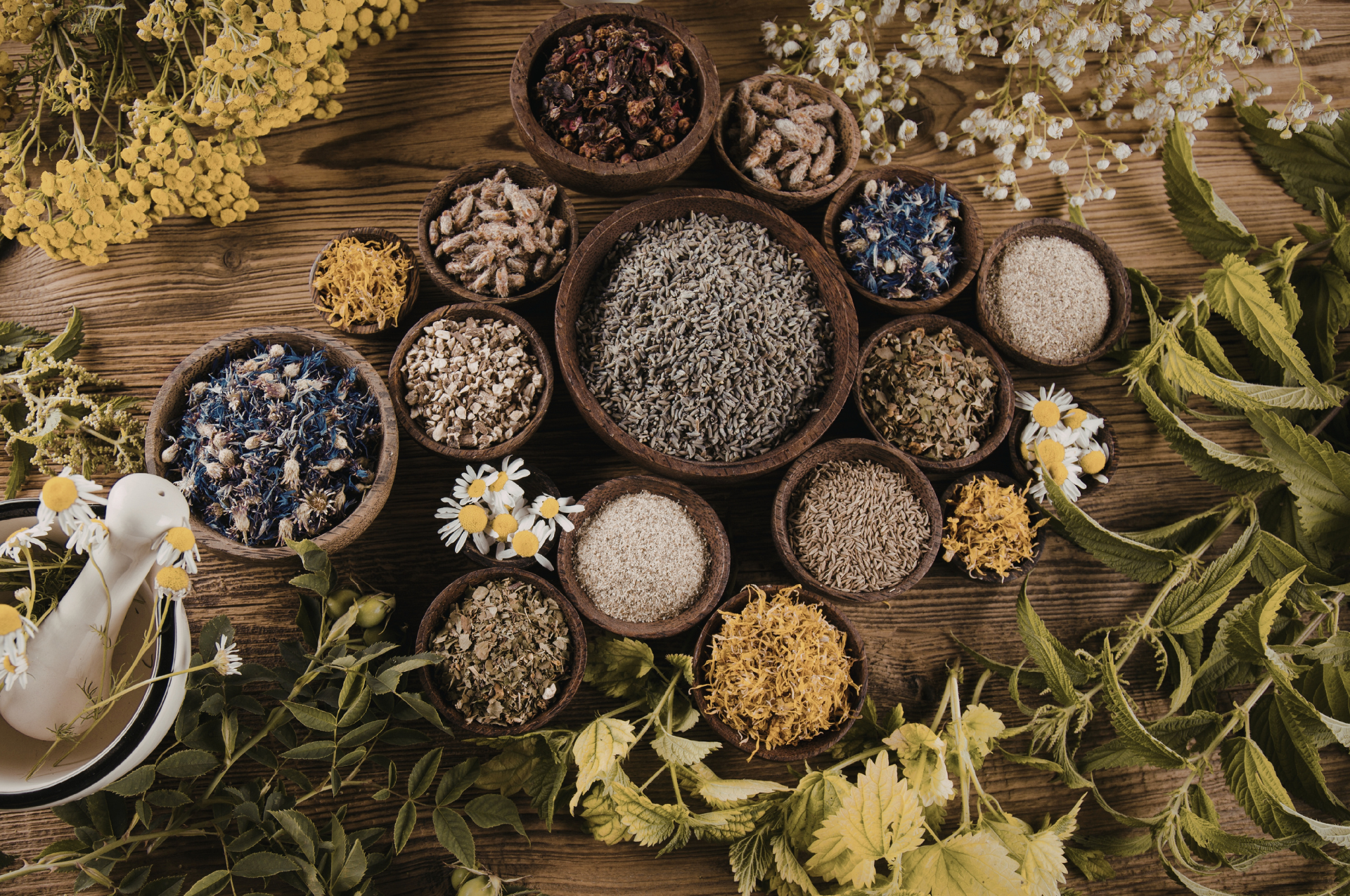 Herbal ingredients in small bowls on a wooden surface with flowers and leaves.